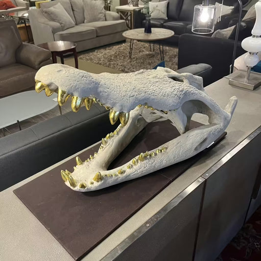 Decorative alligator skull sculpture with gold-painted teeth displayed on a table at Ballard Consignment furniture store.