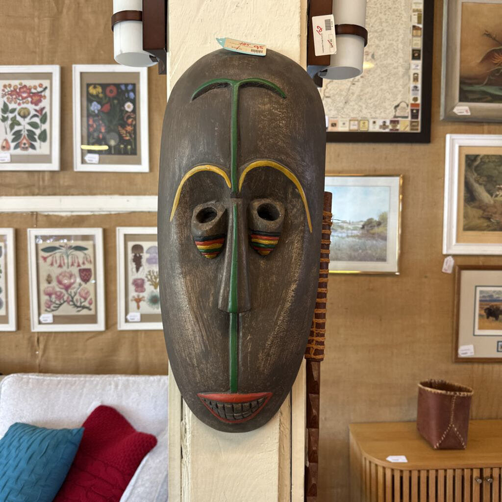 Wooden mask on a stand with framed artwork on the wall in the background