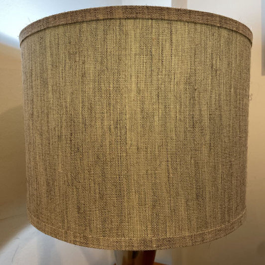 A modern table lamp with a brushed silver hourglass-shaped base and a large cylindrical drum shade made of textured tan linen fabric