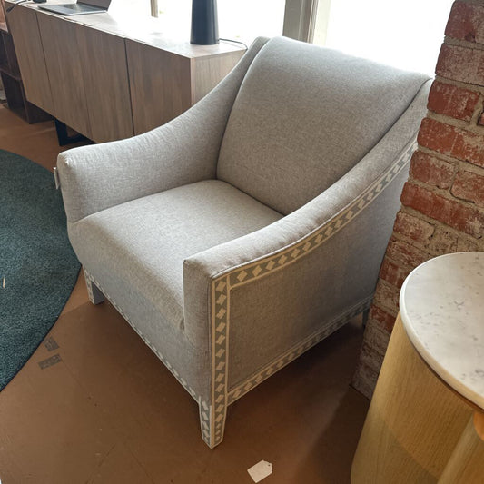 Gray armchair with decorative trim in a room with a brick wall and wooden floor.