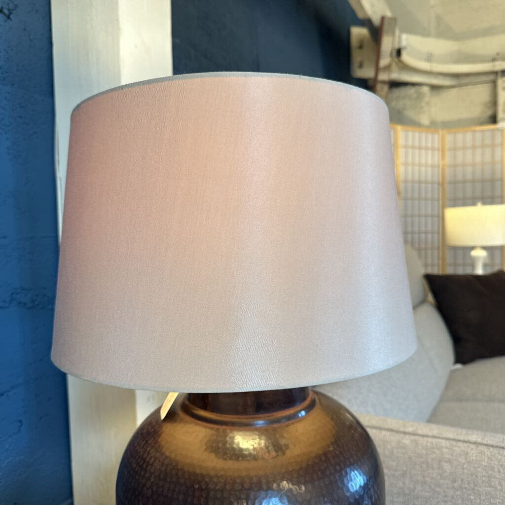 Table lamp with a beige shade on a textured brown base in a room setting.