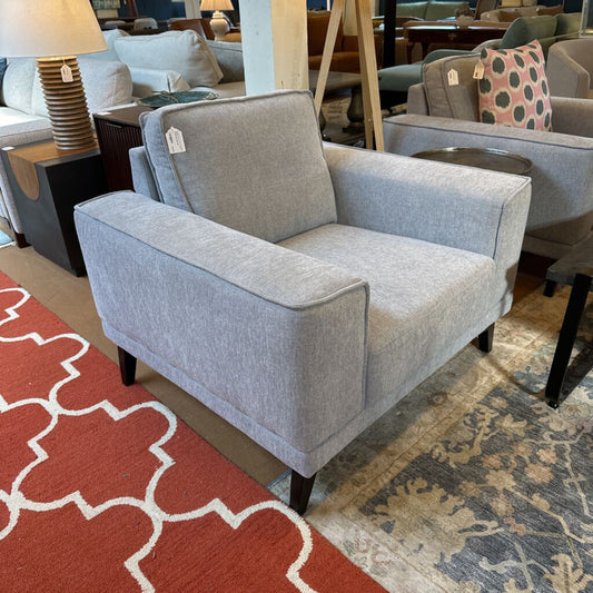 Gray armchair in a showroom with other furniture and a red patterned rug.