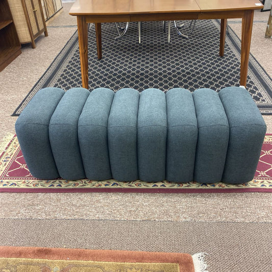 A long grey upholstered bench with a ribbed, modular design consisting of eight rounded vertical segments, resting on a patterned rug