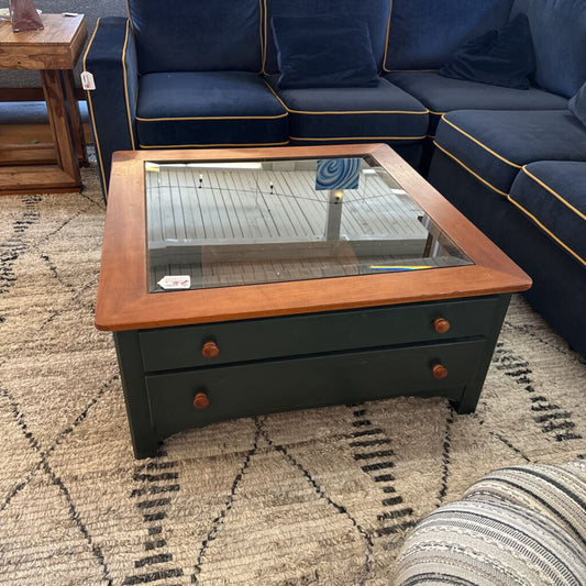 Coffee table with glass top and wooden frame in front of a blue sectional sofa on a patterned rug.