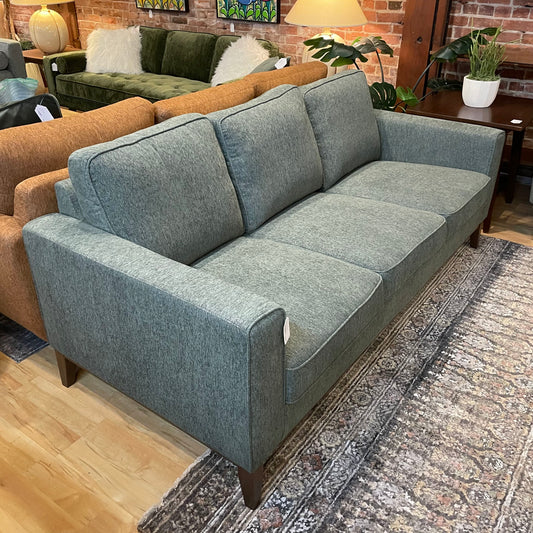 A light blue three-seater sofa with square arms and tapered wooden legs, resting on a dark patterned rug in a furniture showroom