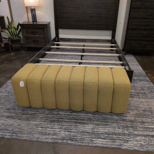 Yellow ottoman in front of a bed with wooden frame on a striped rug.