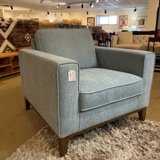 A light blue upholstered armchair with square arms, a single back cushion, and tapered wooden legs resting on a high-pile shag rug