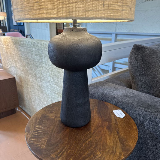 Table lamp with textured base and beige shade on a wooden table in a room setting.