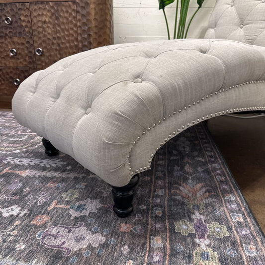 Gray tufted ottoman on a patterned rug with a wooden dresser in the background