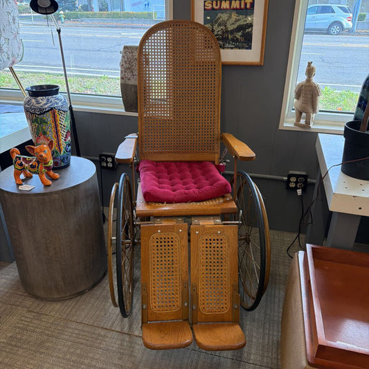 Wooden wheelchair with wicker backrest and pink cushion in a room with windows and various objects.