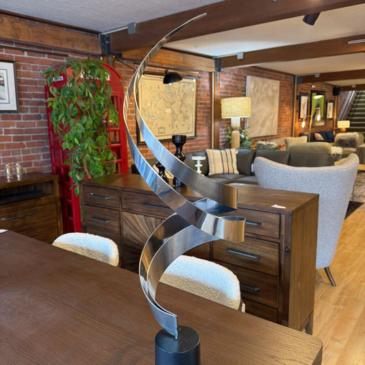 The piece features two wide bands of brushed stainless steel that curve upward in an elegant, interlocking double-spiral design. The sweeping, vertical lines create a sense of weightless energy as they taper toward a single point.