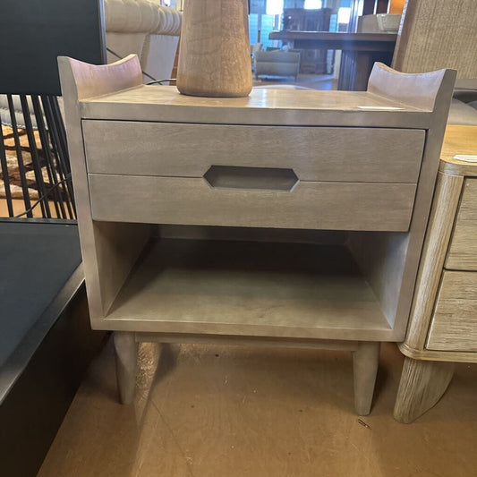 Wooden nightstand with a single drawer and open shelf in a room setting.