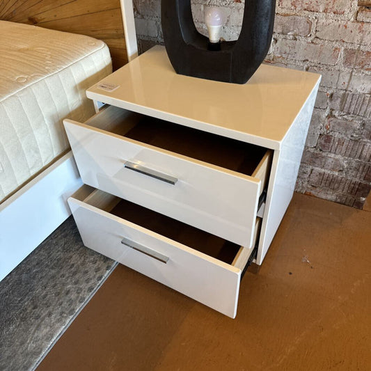 White nightstand with two drawers next to a bed in a room with brick walls.