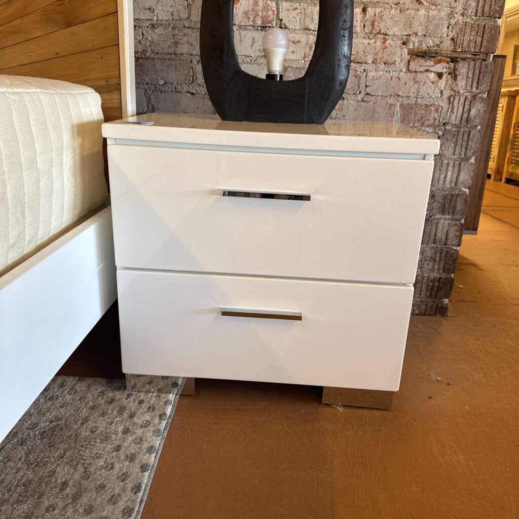 White nightstand with two drawers next to a bed in a bedroom setting.