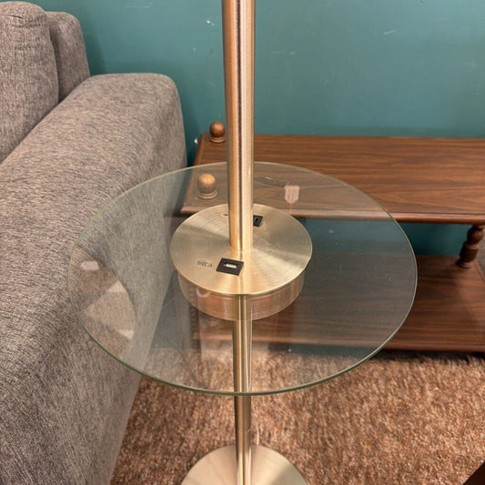 Glass side table with gold metal base next to a gray sofa against a teal wall.