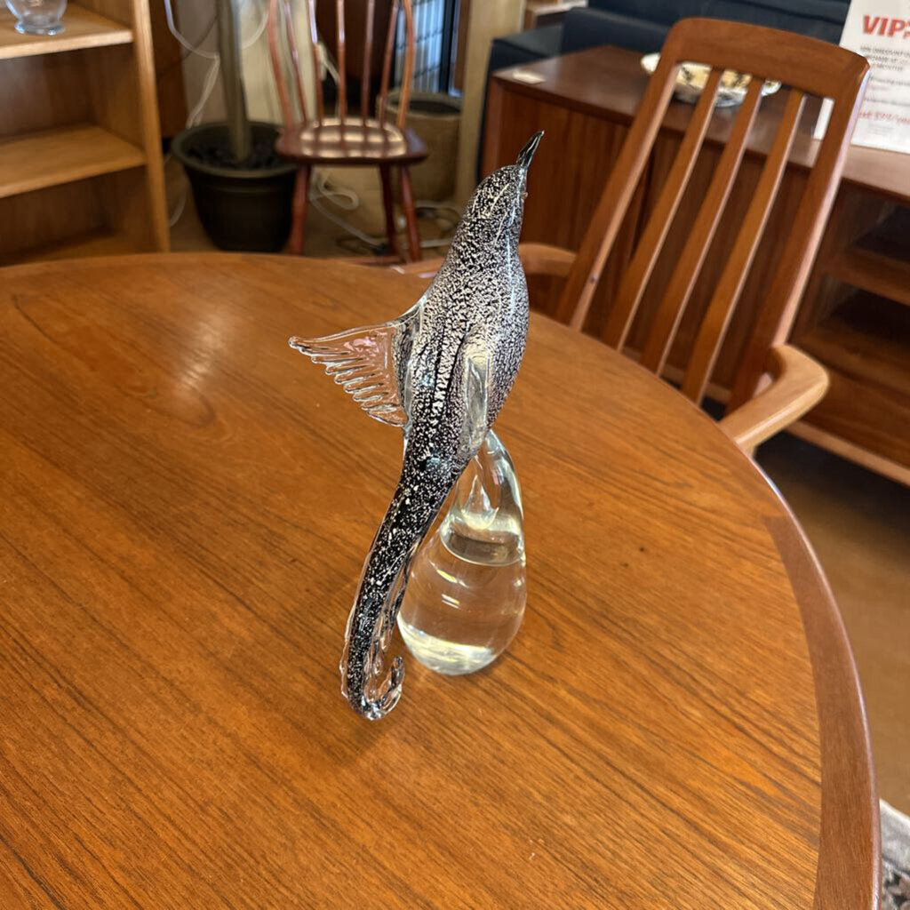 Glass bird figurine on a wooden table with chairs and a bookshelf in the background