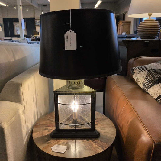 Table lamp with black shade and glass base on a round wooden table in a room setting.