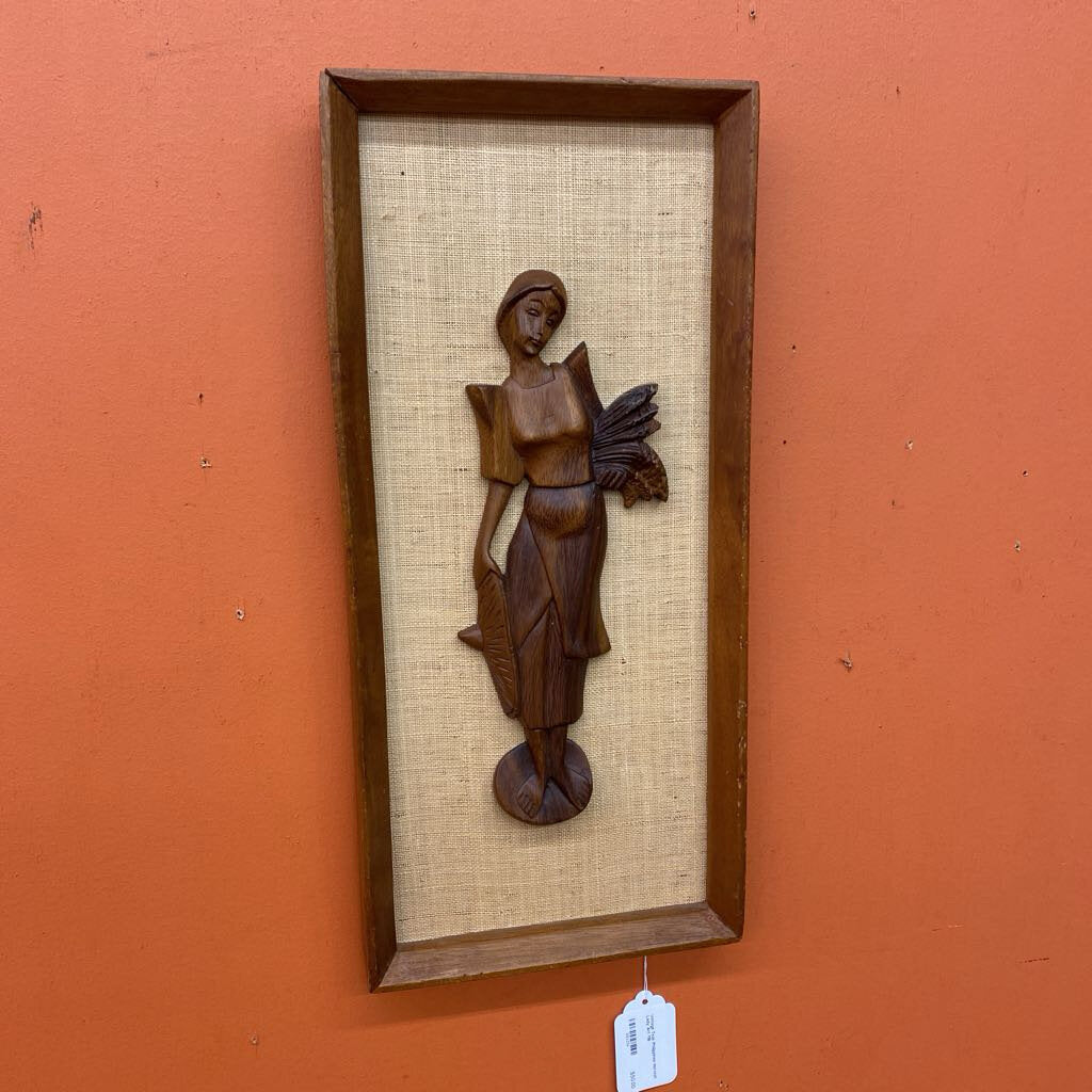 Wooden sculpture of a figure on a textured panel in a wooden frame against an orange wall.