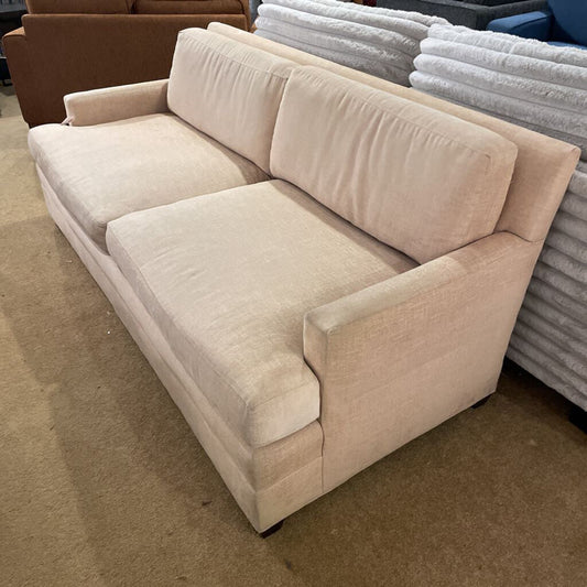 A modern two-cushion sofa upholstered in a tan woven fabric, featuring slim square arms and a low-profile base, shown in a furniture showroom