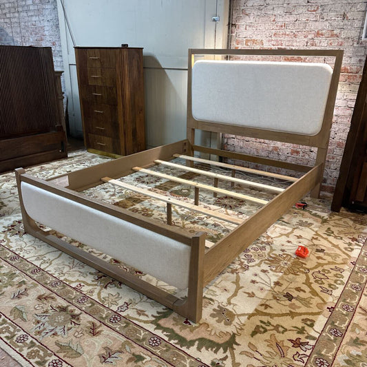 Wooden bed frame with white upholstered headboard on a patterned rug.