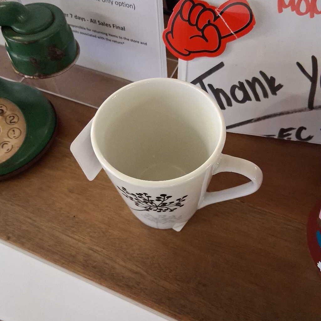 White mug with black design on a wooden surface with a green phone and paper in the background.