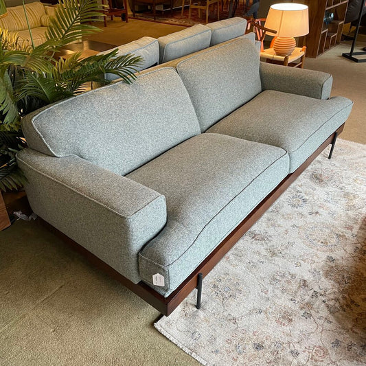 The piece is upholstered in a light seaglass or cool grey textured woven fabric, creating a soft, inviting aesthetic. The sofa features a low-profile frame and a deep two-cushion seat. Its structured silhouette is defined by wide, rectangular track arms and matching plush back cushions.