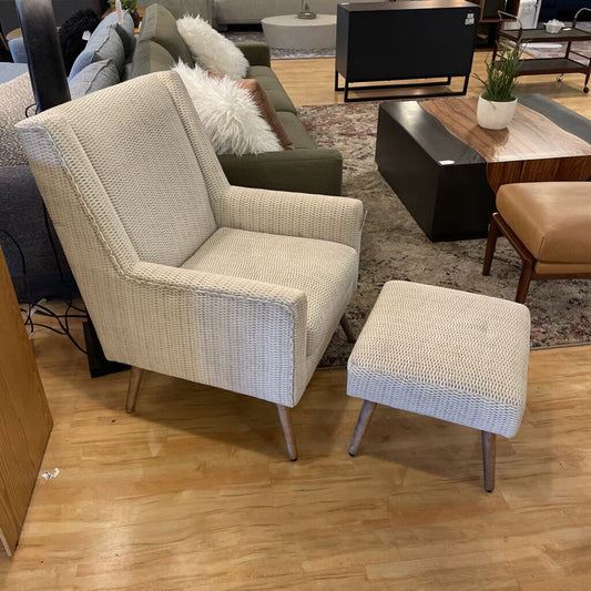 Beige armchair and ottoman on a wooden floor with a living room setting in the background.