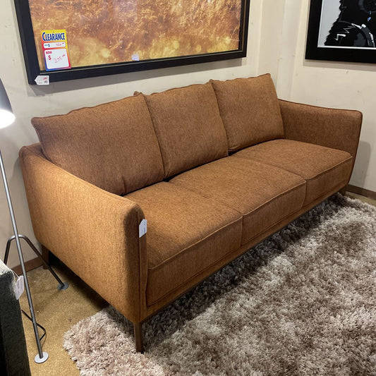 A burnt-orange textured three-seater sofa with three back cushions and a wooden base frame, resting on a high-pile shag rug