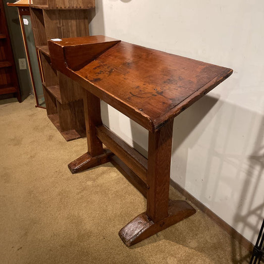 Wooden desk with a slanted top on a beige carpeted floor.