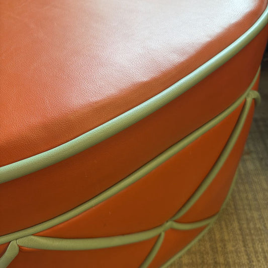 Close-up of an orange leather ottoman with green stitching on a wooden floor.