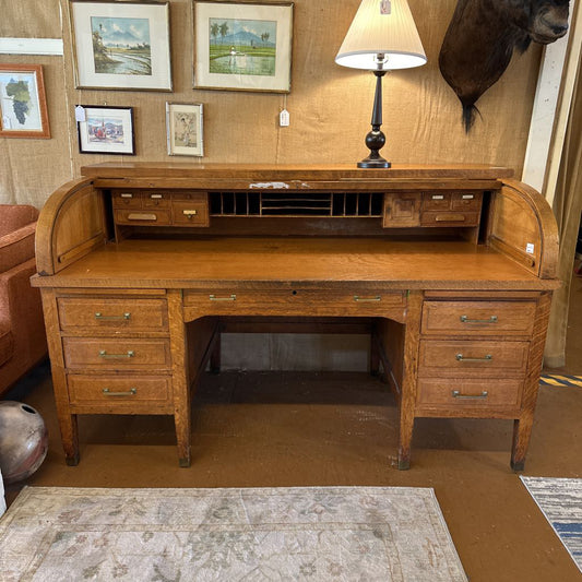 Wooden roll-top desk in a room with framed pictures and a lamp.