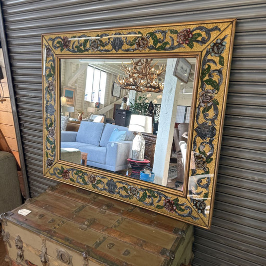 Decorative mirror with floral frame on a wooden chest against a corrugated metal wall.