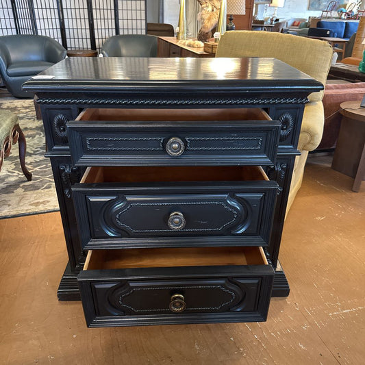 Black three-drawer nightstand in a room with furniture and decor.
