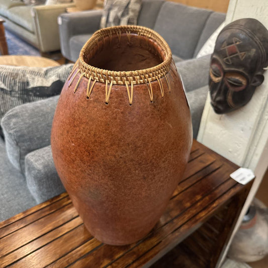 Brown vase with woven top on a wooden table, with a gray couch and decorative mask in the background.