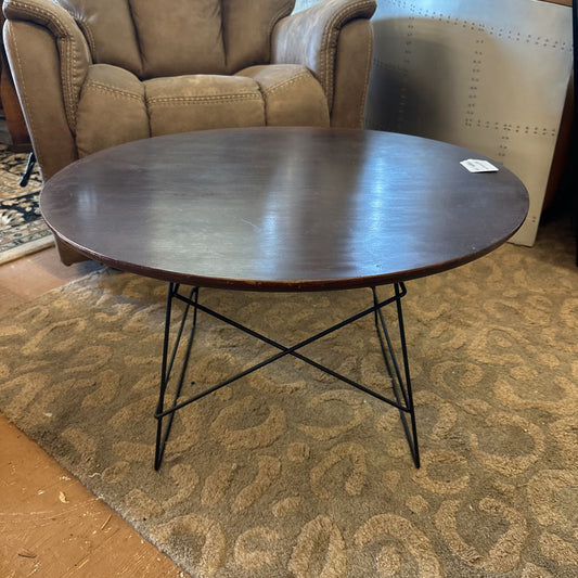 Round wooden table with metal legs on a patterned rug, with a brown leather chair in the background.