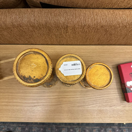 Three gold-colored containers with lids on a wooden surface, one has a label.
