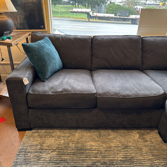 Gray sectional sofa with a teal pillow in a room with a window showing an outdoor view.