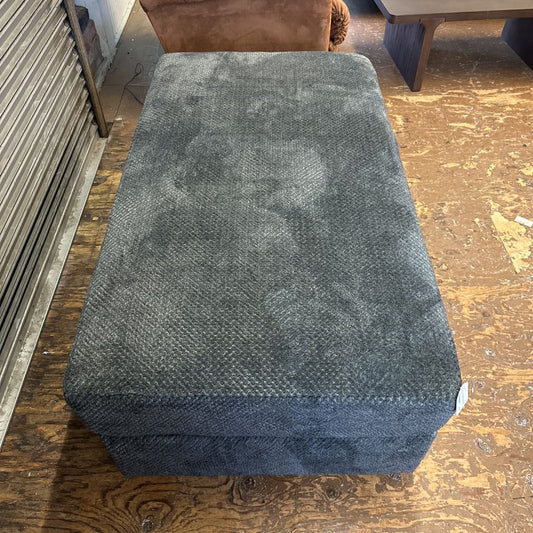 Gray ottoman on a wooden floor with a brown couch in the background