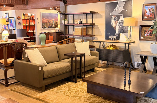 A bright furniture showroom featuring an olive green sofa with white pillows, a dark wood coffee table, and various framed artworks including a large bulldog portrait and a Seattle skyline photo.