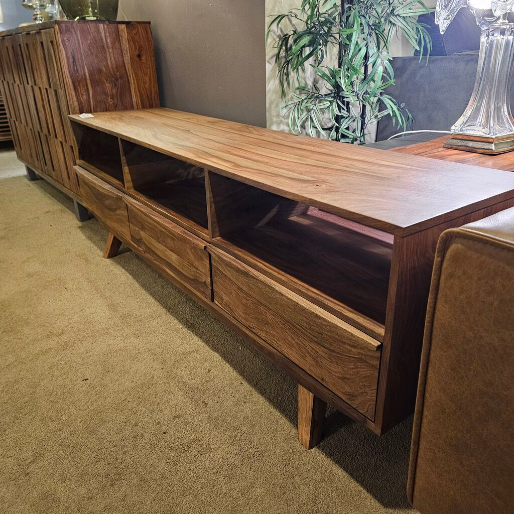 Wooden TV stand in a living room setting with a plant and lamp in the background.