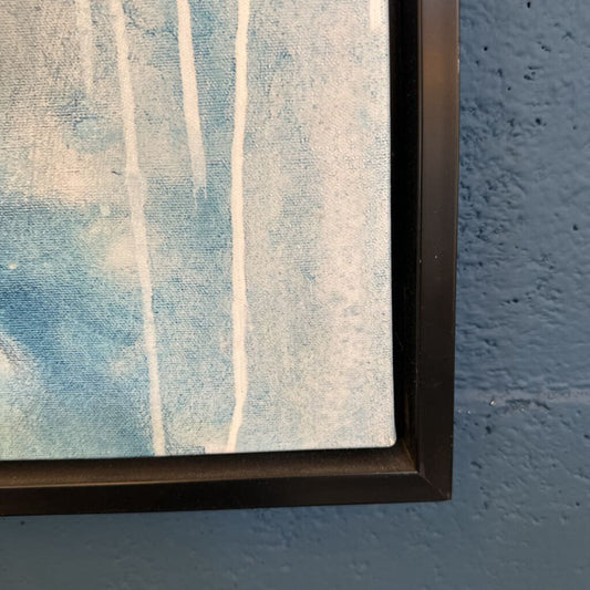 Abstract painting with blue and white strokes in a black frame against a blue wall.