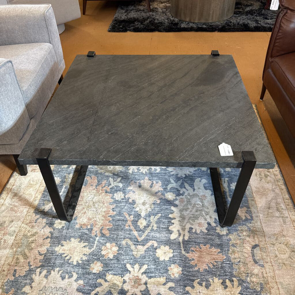 Black marble coffee table with black metal legs on a patterned rug, surrounded by furniture.