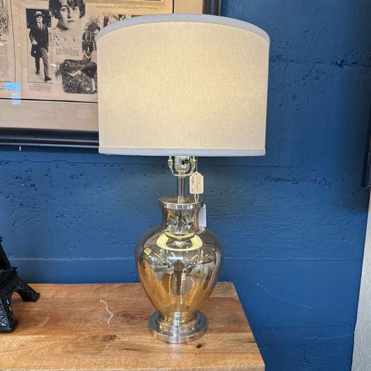 Decorative lamp with a gold base and beige shade on a wooden surface against a blue wall.