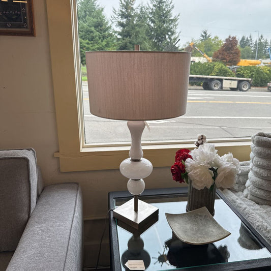 Table lamp with a beige shade on a glass table in a living room with a window view.