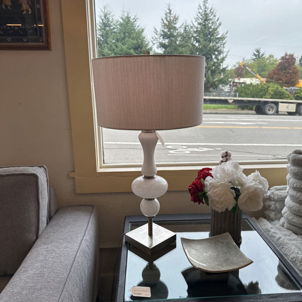Table lamp with a beige shade on a glass table in a living room with a window view.
