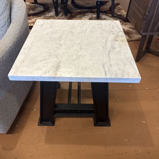 Small square table with marble-like top and black metal legs on a brown floor.