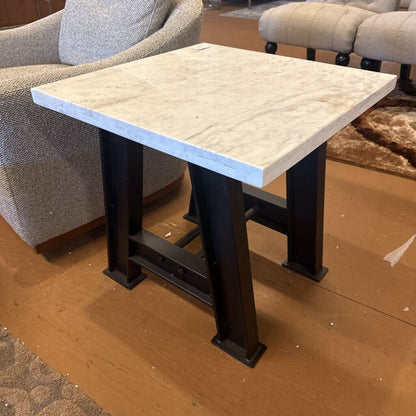 Table with a light-colored top and dark metal legs in a room with furniture.