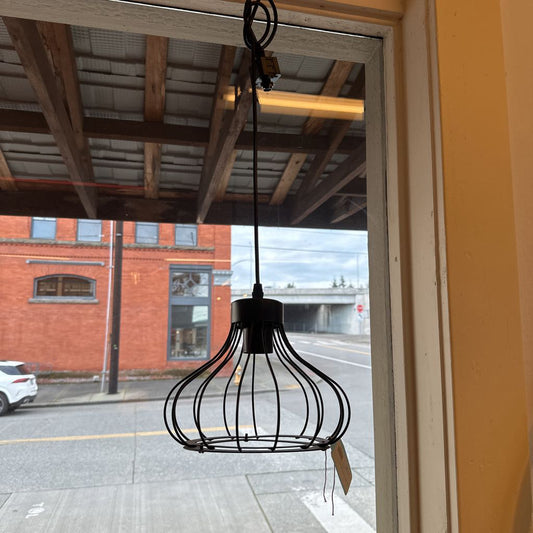 Black wire cage light fixture hanging in front of a window with a view of a street and red brick building.
