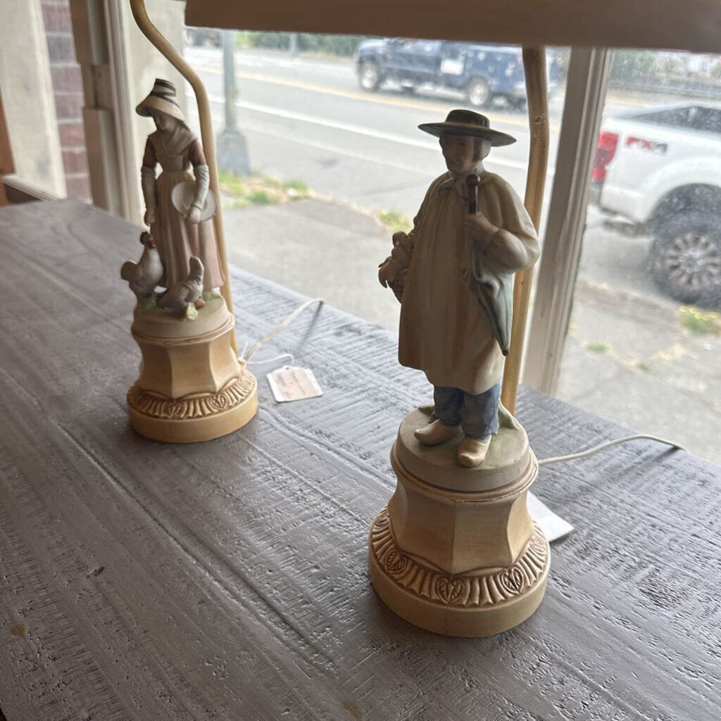 Two decorative lamps with figurines on a table, with a window view of a street.