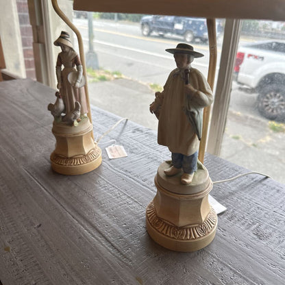 Two decorative lamps with figurines on a table, with a window view of a street.